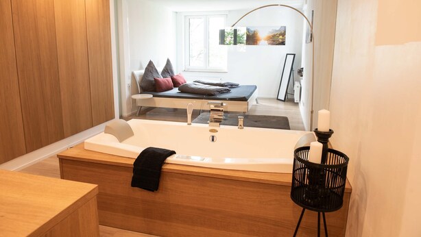 Une salle de bain moderne avec une grande baignoire, un lambris en bois, un porte-bougie noir et une vue sur une chambre à coucher avec un lit et des oreillers.