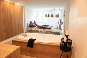 A modern bathroom with a large bathtub, wooden paneling, a black candle holder, and a view of a bedroom with a bed and pillows. - The City Suite - Loft 4 (Kempten (Allgäu))