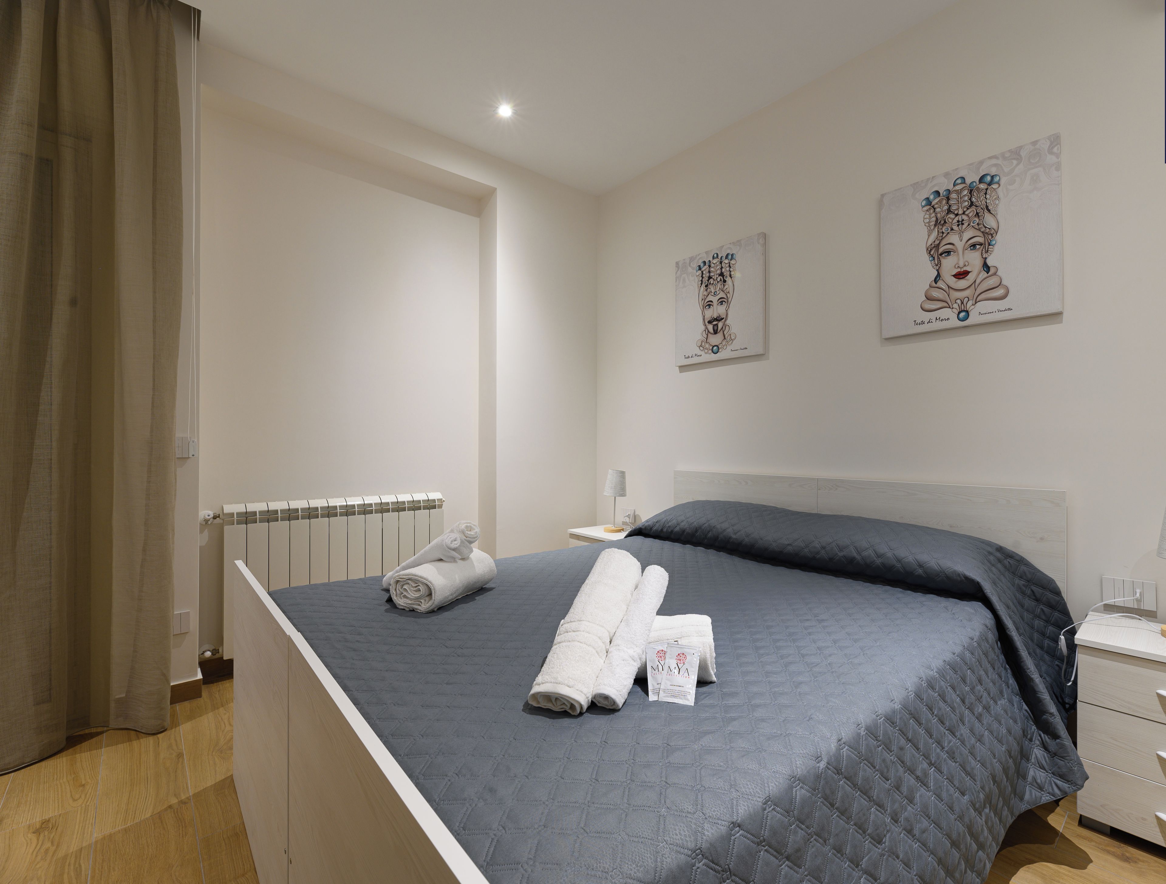 A neatly made bed with rolled towels, a bedside table with a lamp, and two artworks on the wall.