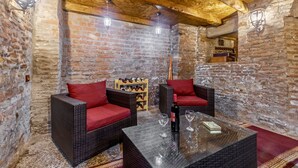 Une pièce rustique aux murs en briques apparentes, avec des poutres en bois, des meubles en osier agrémentés de coussins rouges, et une bouteille de vin posée sur une table en verre.