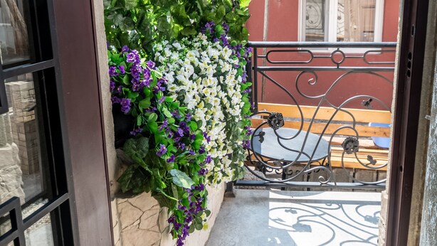 Une porte-fenêtre donnant sur un balcon avec un mur en pierre, une rambarde en métal et une vue sur un bâtiment rouge avec un toit en tuiles.