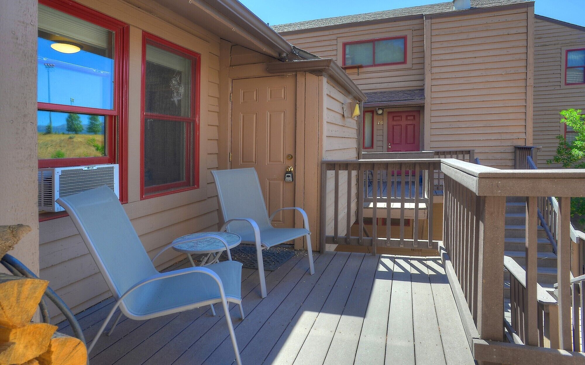 A wooden deck with two chairs and a small table, a red door, and a view of a neighboring house.