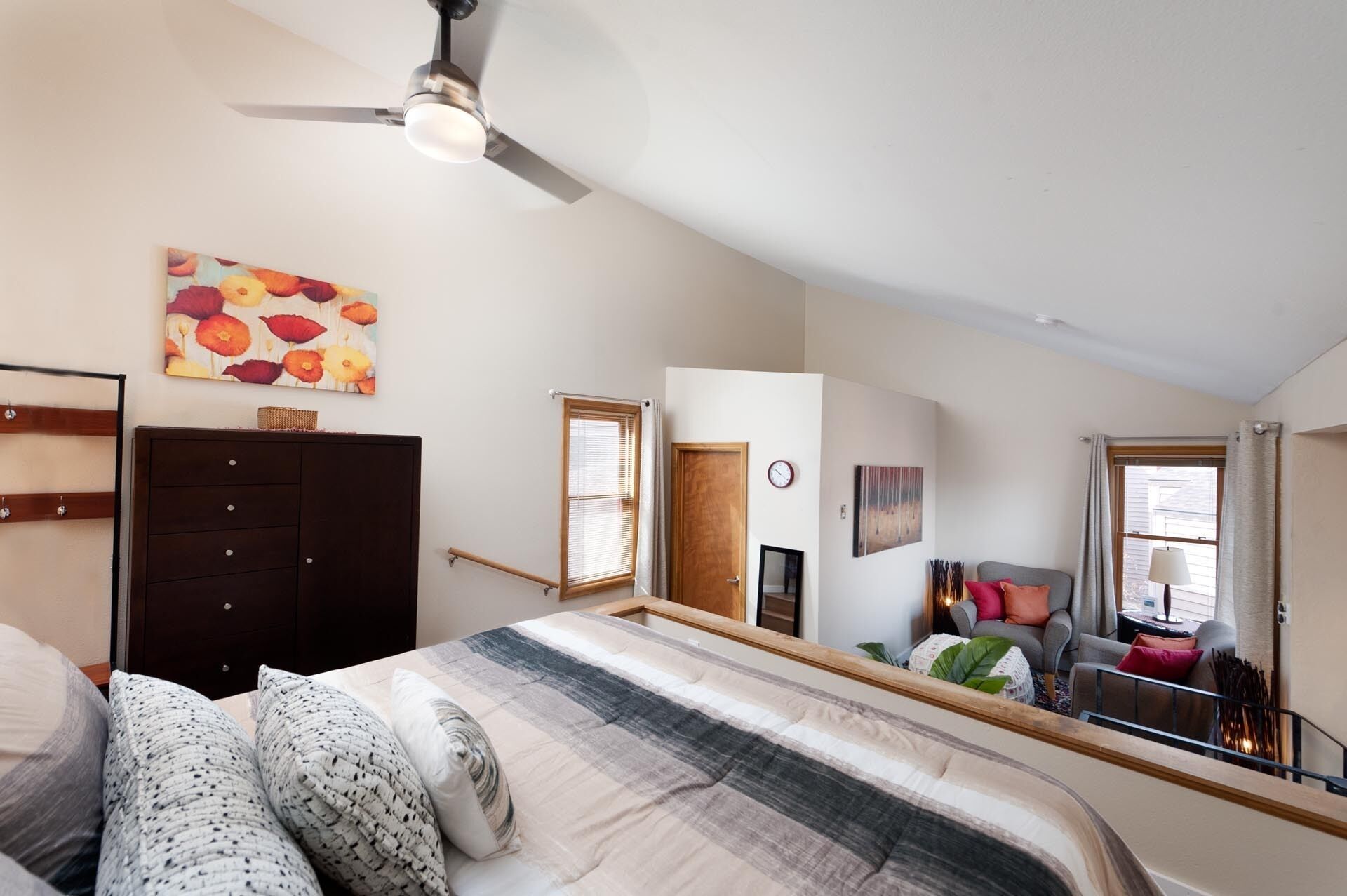 A bedroom with a bed, a dresser, a ceiling fan, a painting, a window, and a door.