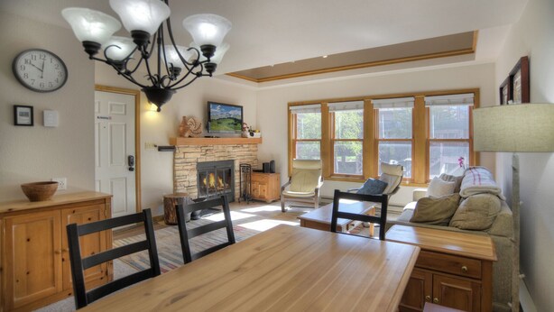 A cozy living room with a fireplace, wooden furniture, and a large window with blinds. - Village View Vista | Walk to Town, Pool/Hot Tub - Near FLC, Golf (Durango)