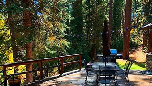 Une terrasse en bois avec une table et des chaises, entourée de grands arbres et d’un mur de pierre.