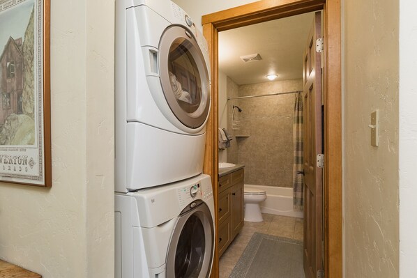 Ein Waschbereich mit einer Frontlader-Waschmaschine und einem Trockner, ein Badezimmer mit Dusche, Toilette und Waschbecken sowie eine Holztür zum Duschbereich.