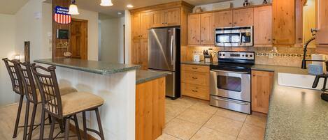 A kitchen with wooden cabinets, a stainless steel refrigerator, an oven, and a microwave. There are bar stools and a countertop.