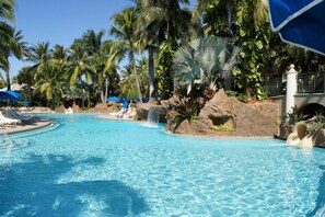 Ein klares, blaues Schwimmbecken, umgeben von Palmen und Liegestühlen.