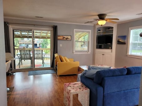 A living room with a blue sofa, yellow armchair, and a television. There is a sliding glass door leading to an outdoor area with a deck.