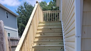 Una escalera de madera que sube a un área de terraza, rodeada por una valla de madera y una casa con revestimiento.