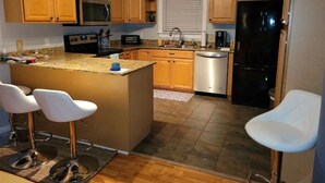 Cocina con armarios de madera, encimera de granito, refrigerador de acero inoxidable y dos taburetes blancos de bar.
