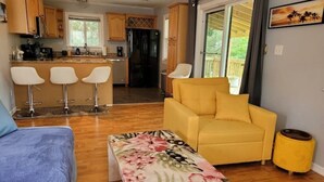 Un espacio habitable compacto con sofá, una pequeña mesa con mantel decorado con motivos florales, un sillón amarillo, y una zona de cocina con armarios de madera y un refrigerador.