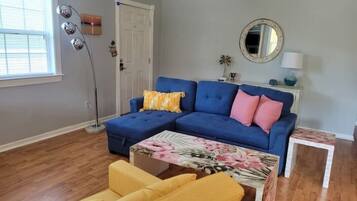 A living room with a blue sectional sofa, a yellow armchair, a floral coffee table, a floor lamp, and a round mirror on the wall.