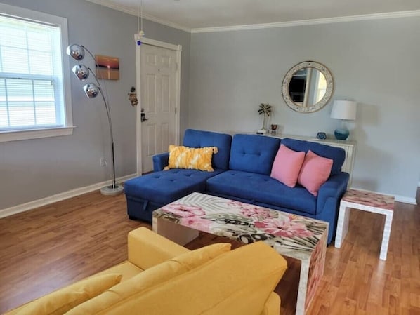 Een woonkamer met een blauwe modulaire bank, een gele fauteuil, een bijzettafeltje met bloemenprint, een vloerlamp en een ronde spiegel aan de muur.