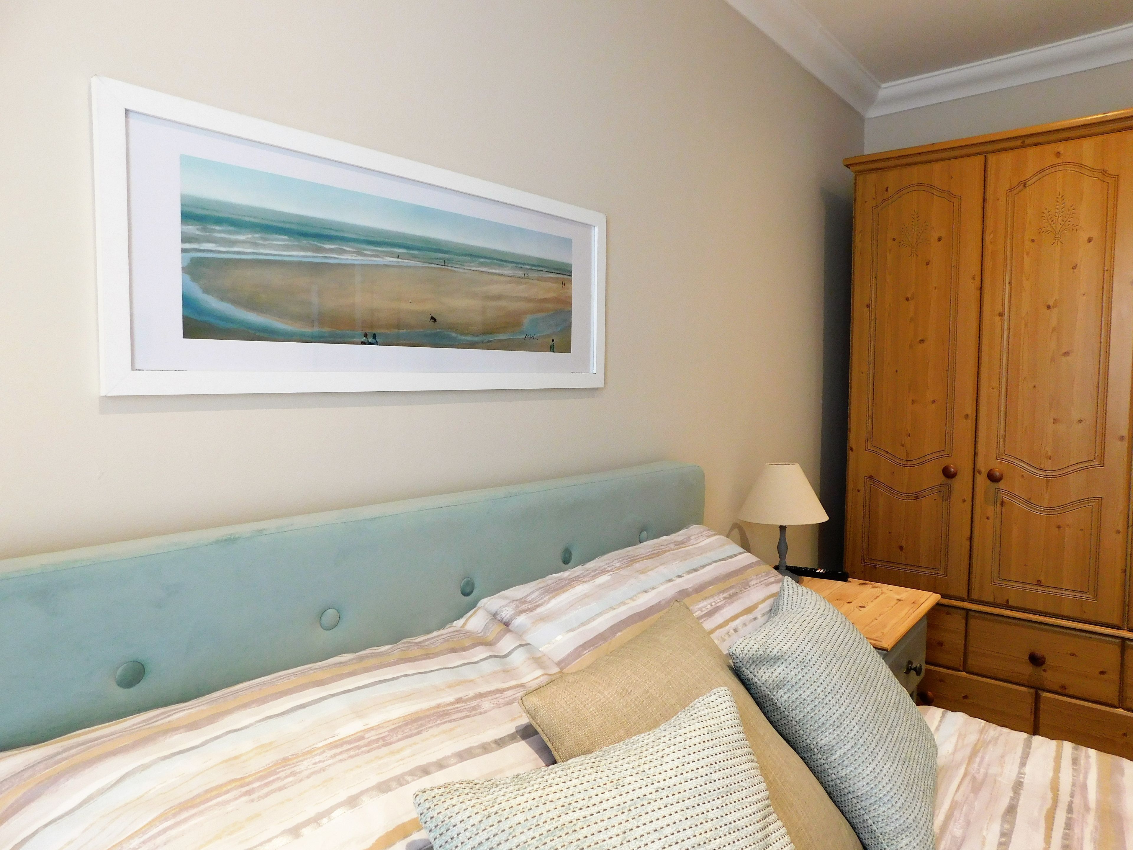 A bedroom with a bed, a framed landscape painting, a wooden wardrobe, and a bedside table with a lamp.