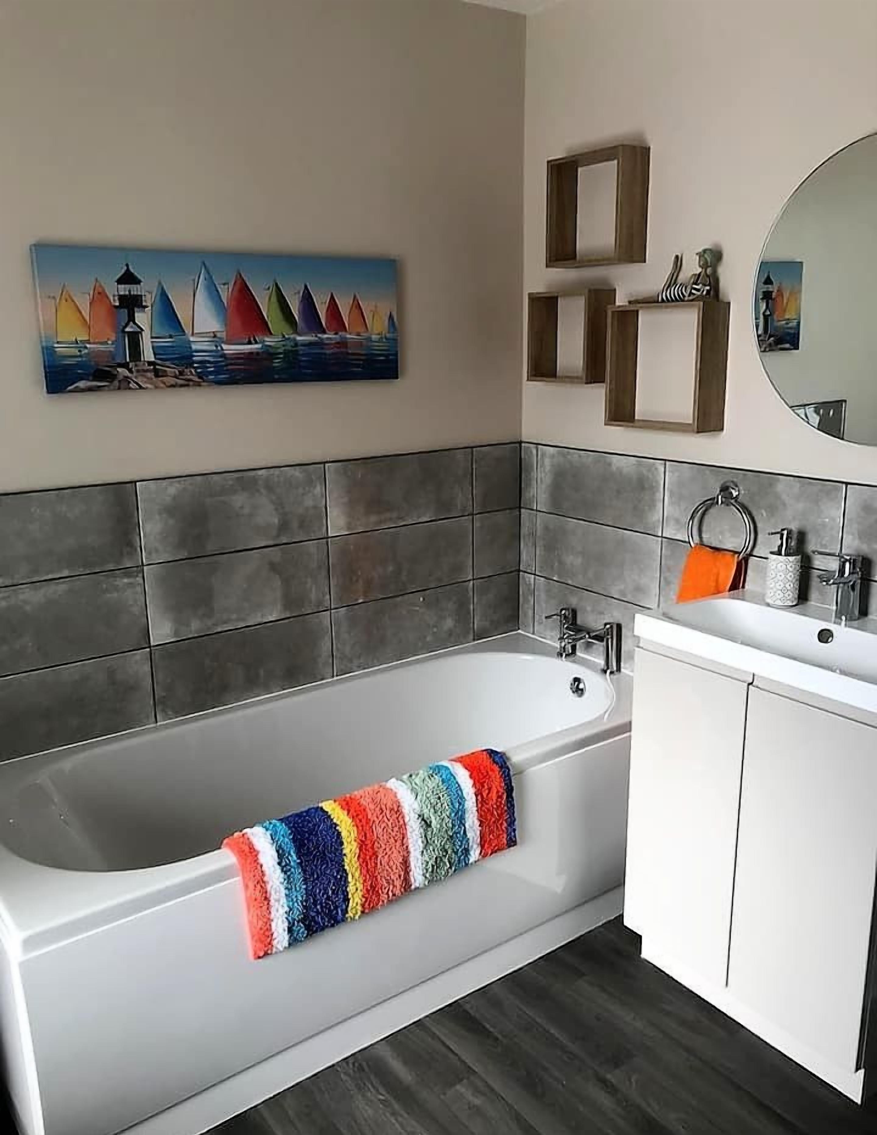 Een moderne badkamer met een groot bad, grijze betegelde wanden, een kleurrijke handdoek, een wastafel met spiegel en een schilderij van zeilboten.