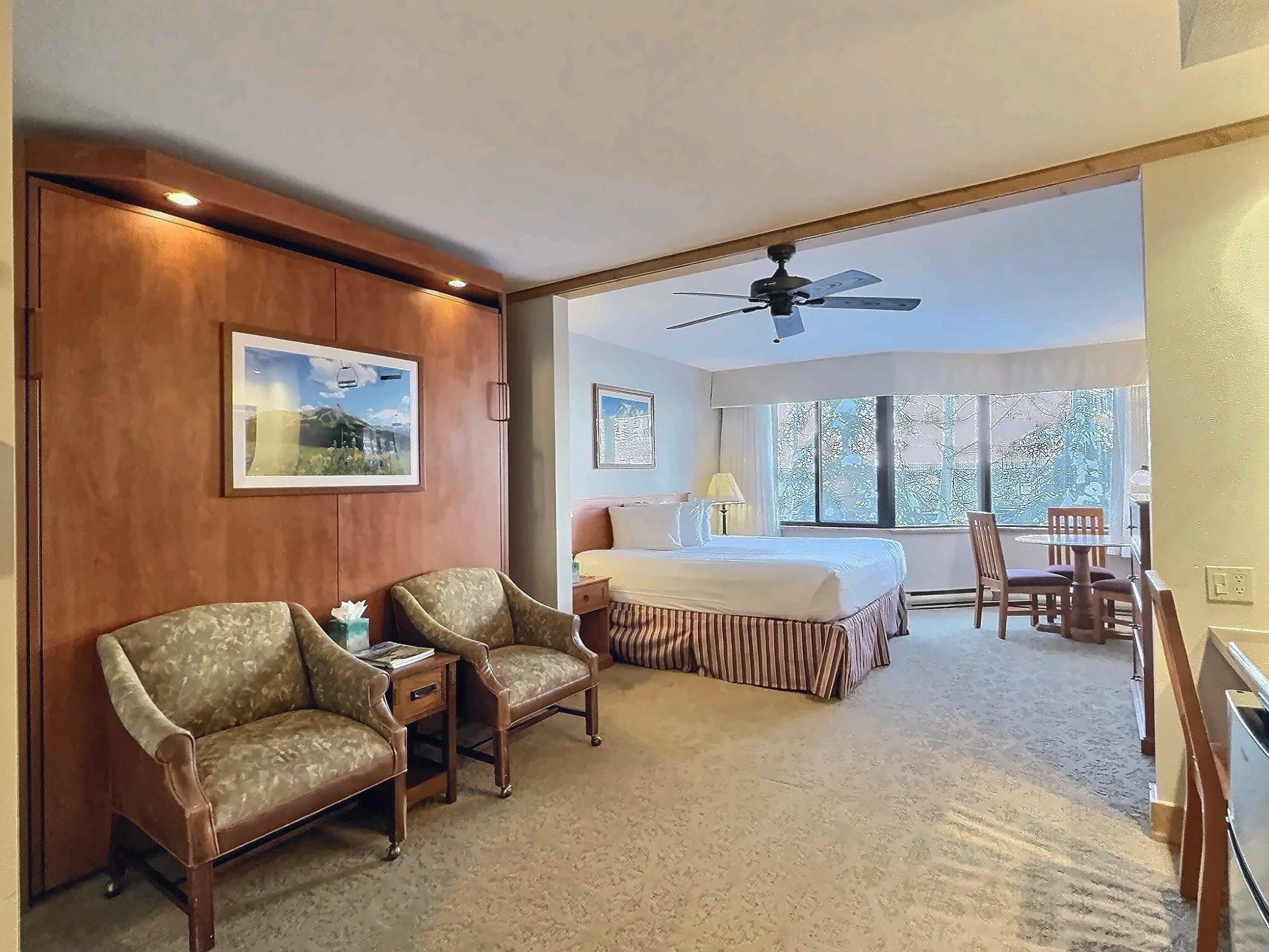 Une chambre d’hôtel comprenant un lit, deux fauteuils, un bureau et une table à manger. Elle est dotée de deux fenêtres qui laissent entrer la lumière naturelle et d’un ventilateur de plafond.