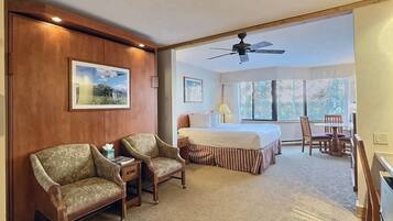 Une chambre d’hôtel comprenant un lit, deux fauteuils, un bureau et une table à manger. Elle est dotée de deux fenêtres qui laissent entrer la lumière naturelle et d’un ventilateur de plafond.