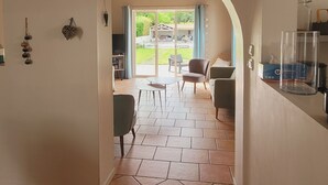 Un couloir avec une grande horloge, un sol carrelé et, au fond, une vue sur un espace extérieur accessible par une arche, où se trouvent une table et des chaises.
