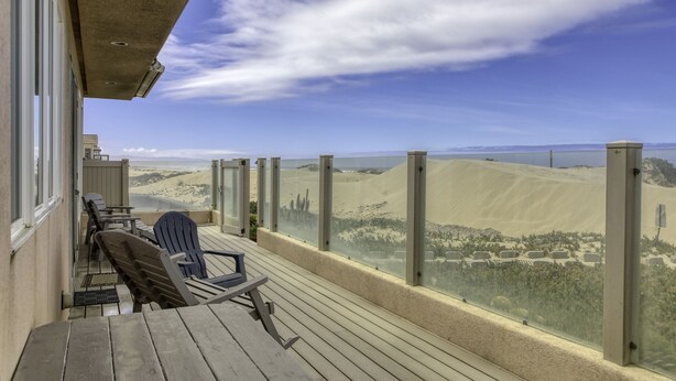 Una terraza de madera con mobiliario exterior, con vistas a un paisaje desértico de dunas de arena y una masa de agua.