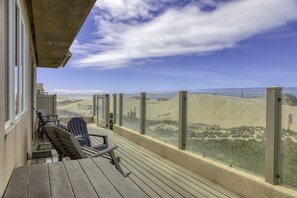 Una terraza de madera con mobiliario exterior, con vistas a un paisaje desértico de dunas de arena y una masa de agua.