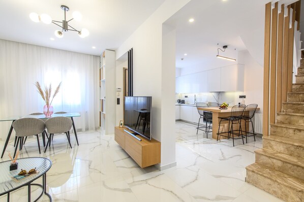 Una sala moderna con televisor, una mesa de comedor y sillas, una cocina con taburetes de bar, y una escalera.