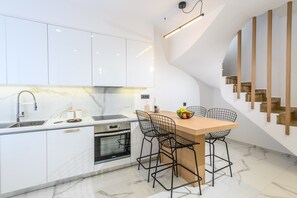 Una cocina moderna con armarios blancos, encimera de mármol, horno empotrado y mesa de comedor de madera con sillas de alambre.