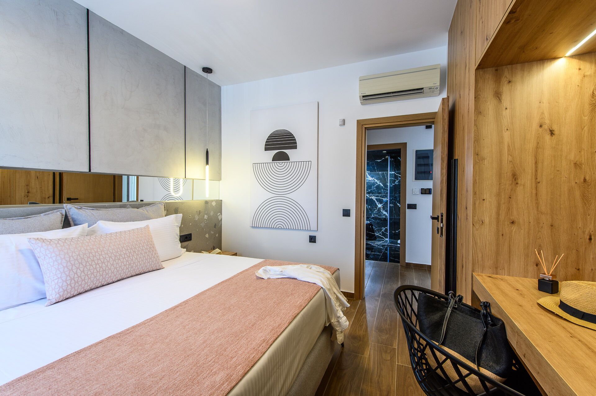 Ein modernes Schlafzimmer mit einem großen Bett, einem Holztisch und einem Stuhl. Eine Tür führt in einen anderen Raum, und an der Wand ist eine Klimaanlage angebracht.