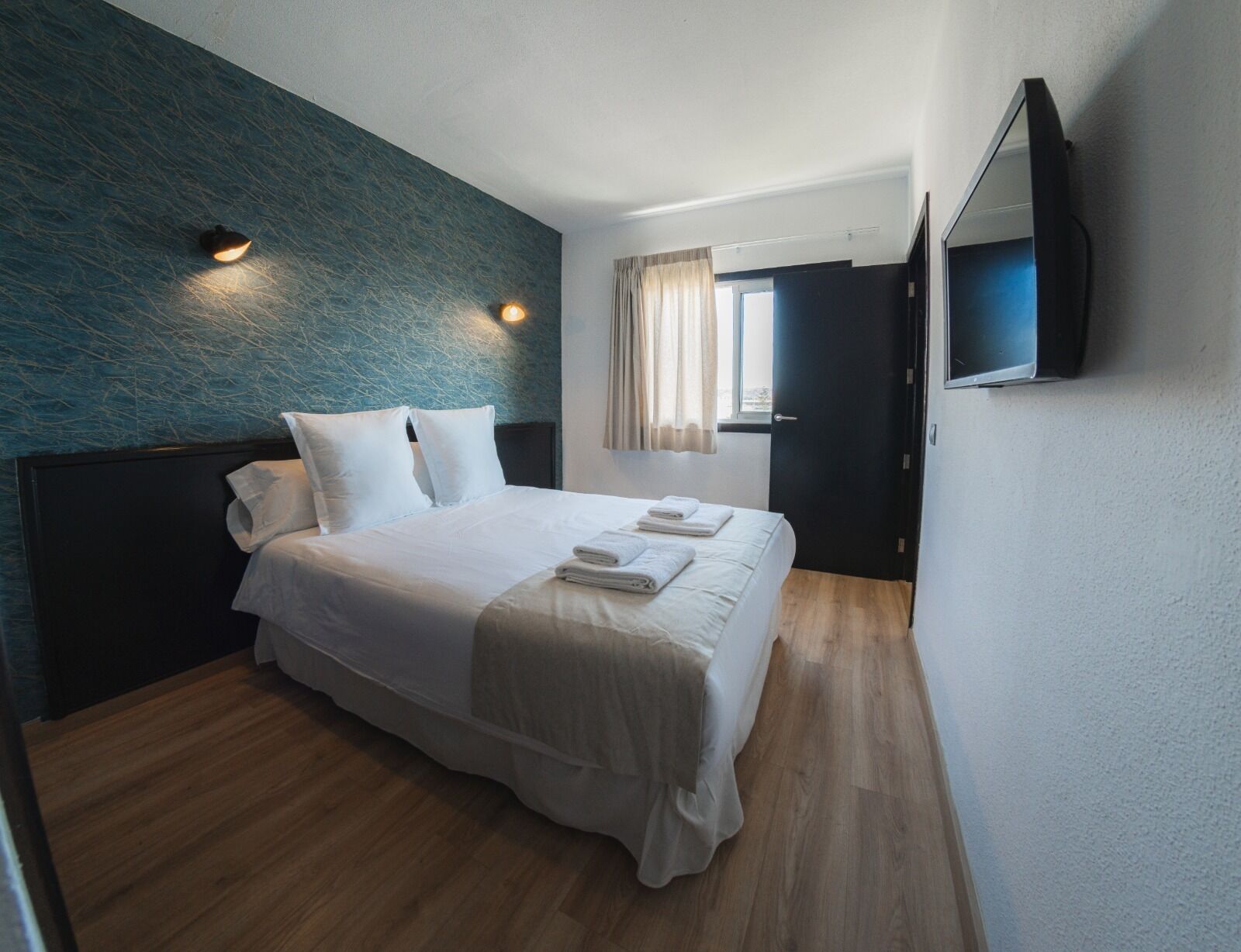 A hotel room with a bed, a flat-screen TV mounted on the wall, and a window with curtains.