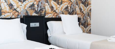 Habitación de hotel con dos camas, ropa de cama blanca, papel tapiz con motivos florales y una mesita con toallas y artículos de aseo.