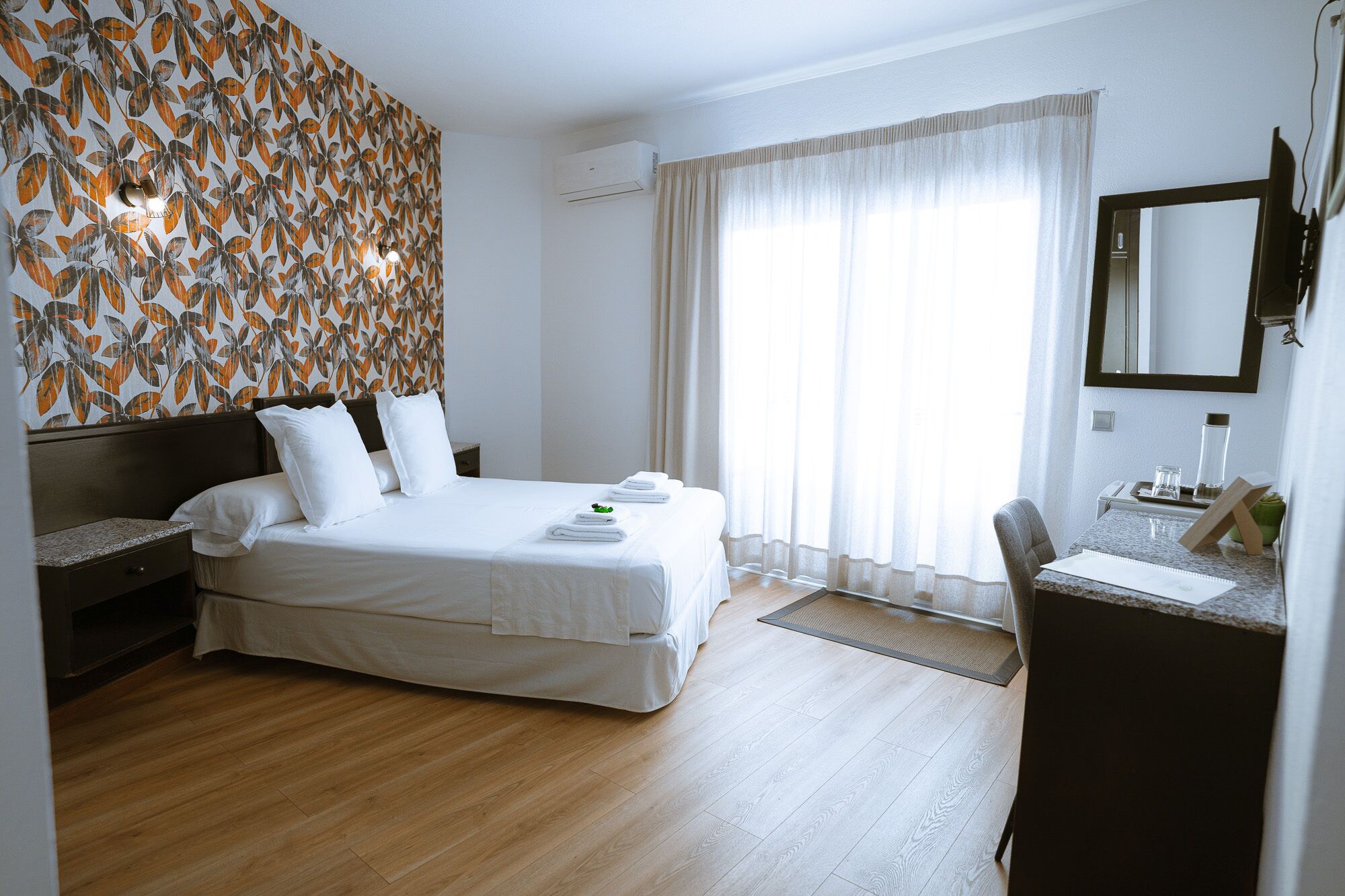 Una habitación de hotel con una cama grande, un escritorio con silla, un espejo y una ventana con cortinas.