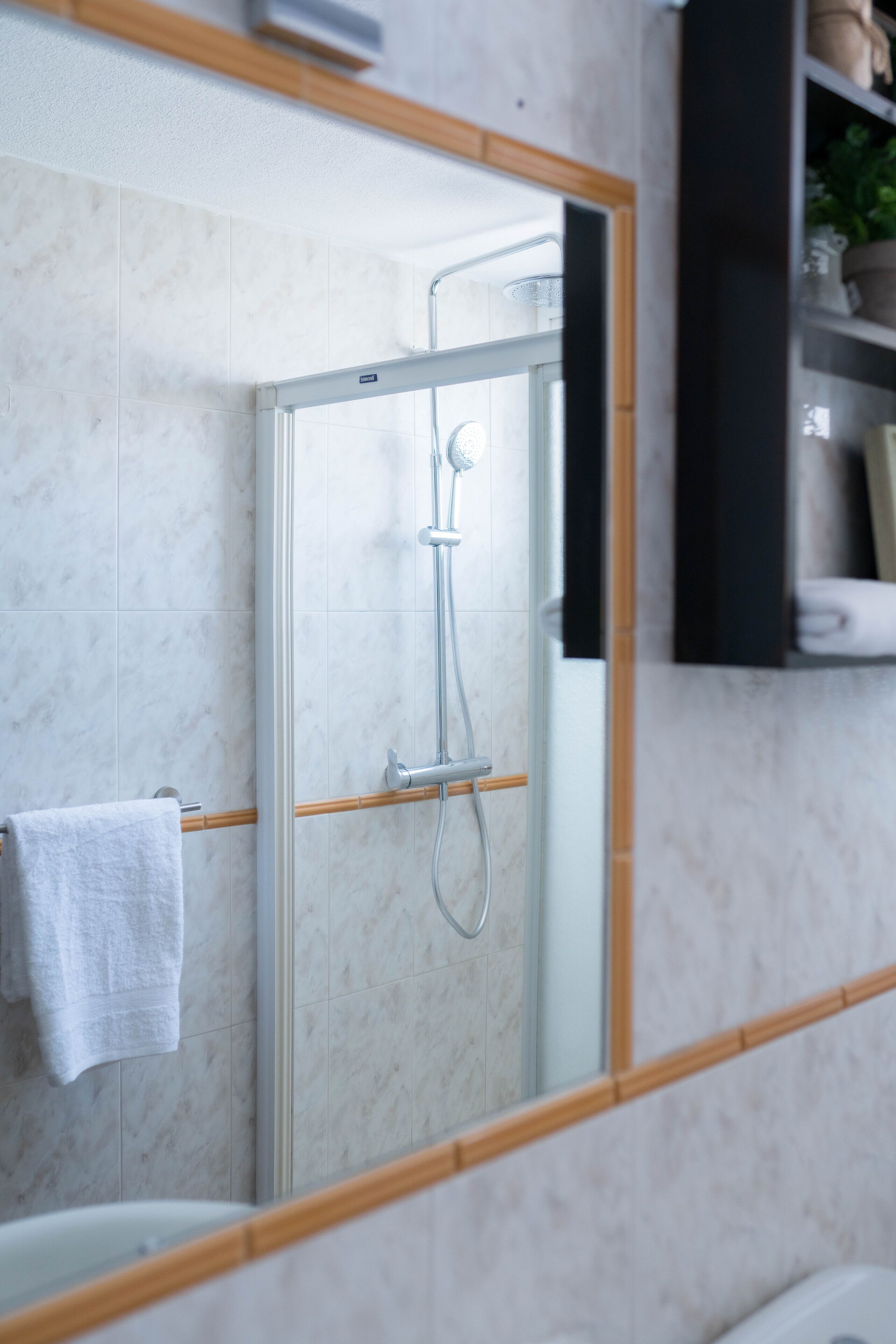A modern bathroom with a glass-enclosed shower, a white towel hanging on the wall, and a wooden shelf with toiletries.