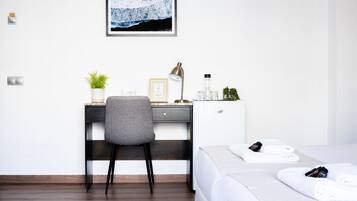 A hotel room with a bed, a desk with a chair, a framed picture on the wall, and a small potted plant.