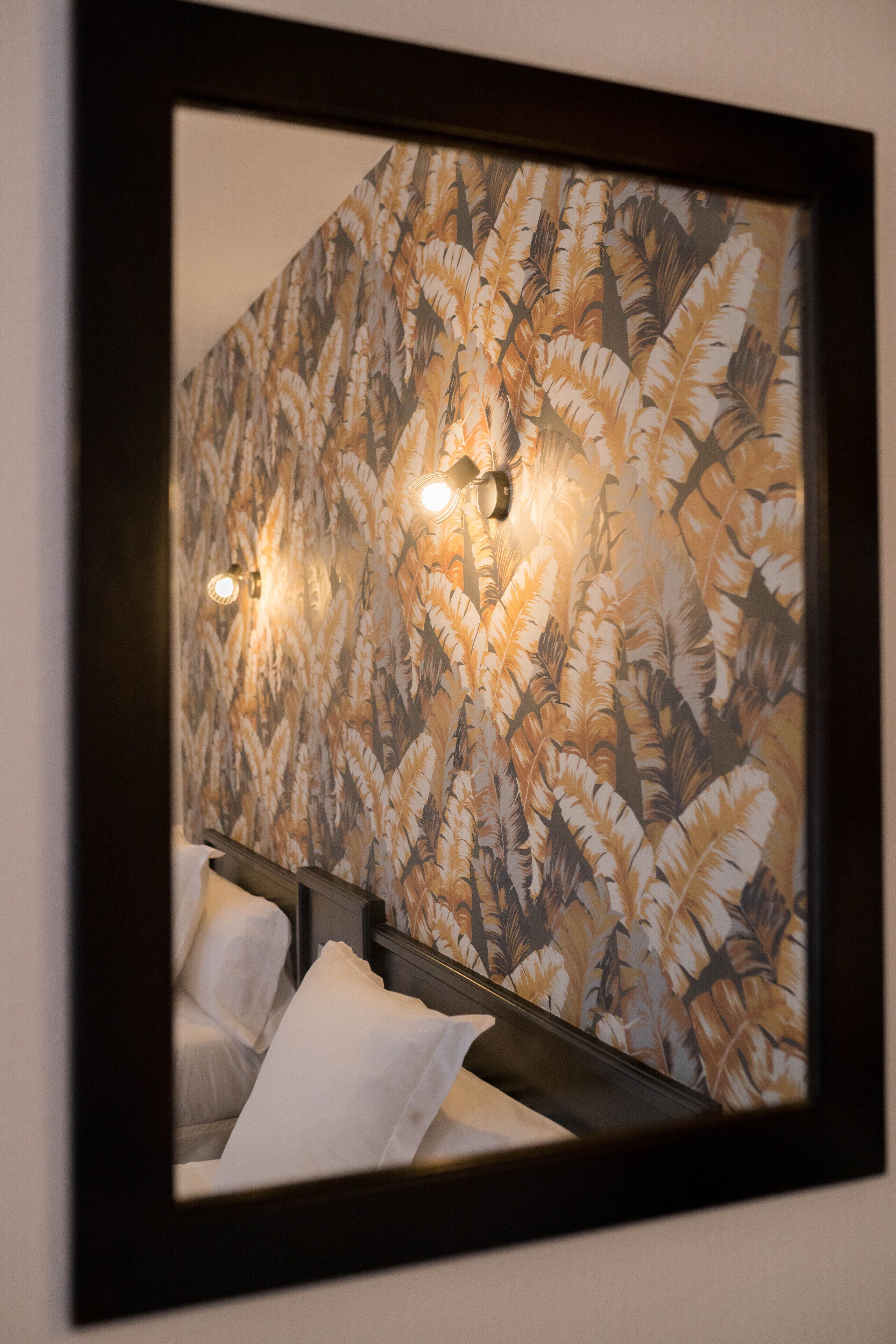 A hotel room with a large mirror reflecting a wall with a feather pattern. There are two bedside lamps and a bed with white linens.
