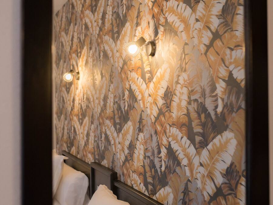 Quarto de hotel com um espelho grande refletindo uma parede com padrão de penas. Há duas luminárias de cabeceira e uma cama com roupa de cama branca.
