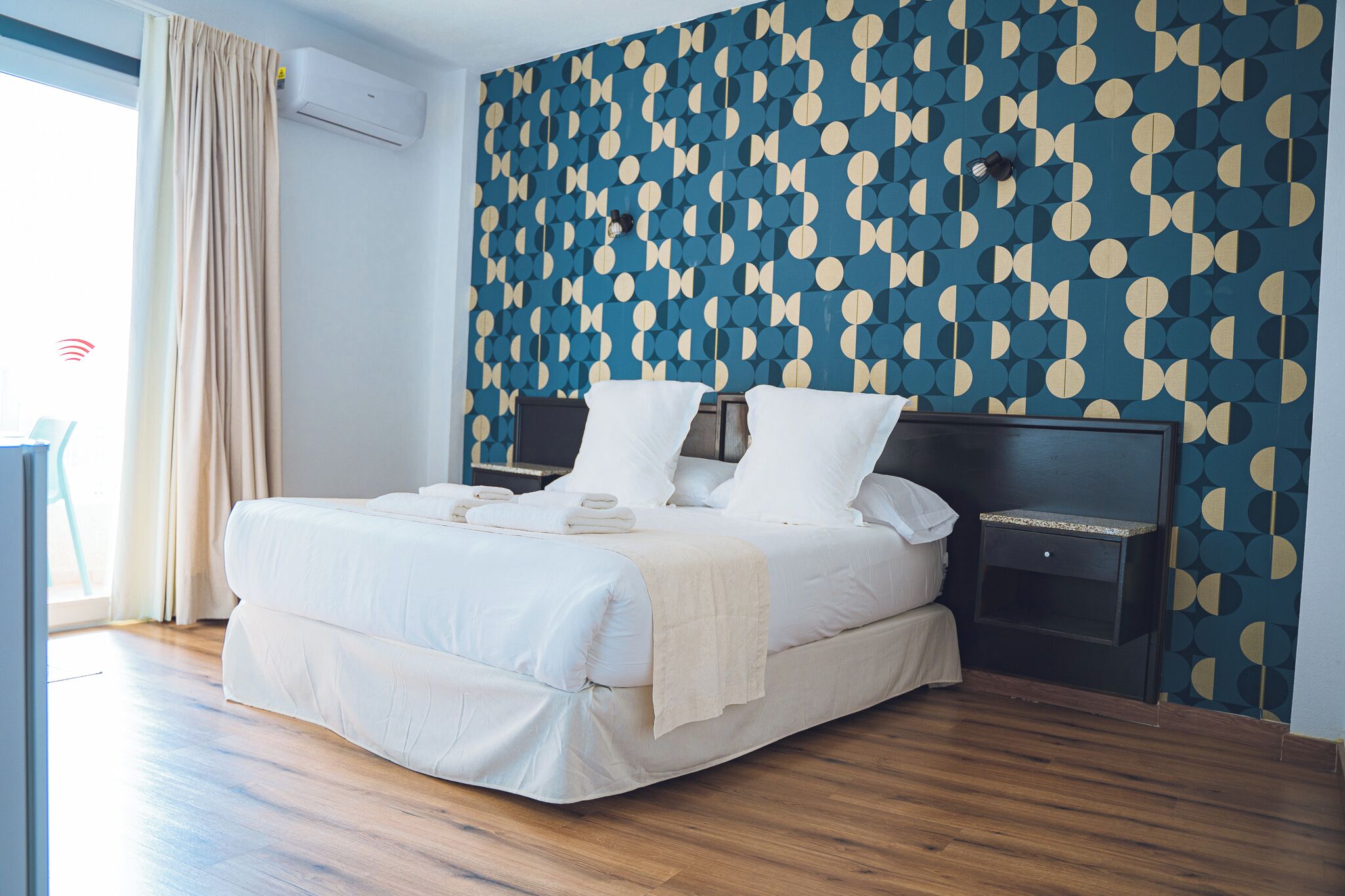 A neatly made bed with white linens and pillows, a bedside table with a lamp, and a wall with a geometric pattern.