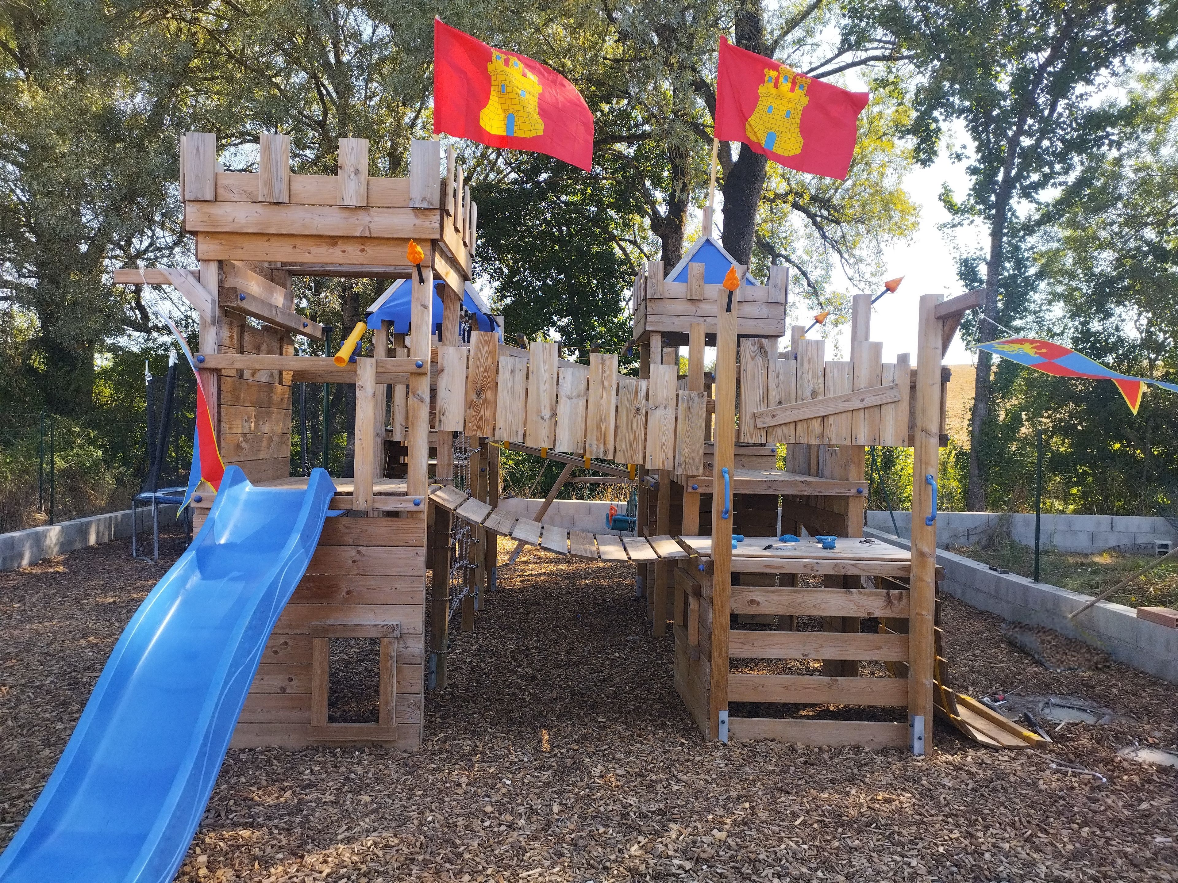 Une structure de jeux en bois, dotée d’un toboggan bleu, d’éléments d’escalade et de drapeaux.