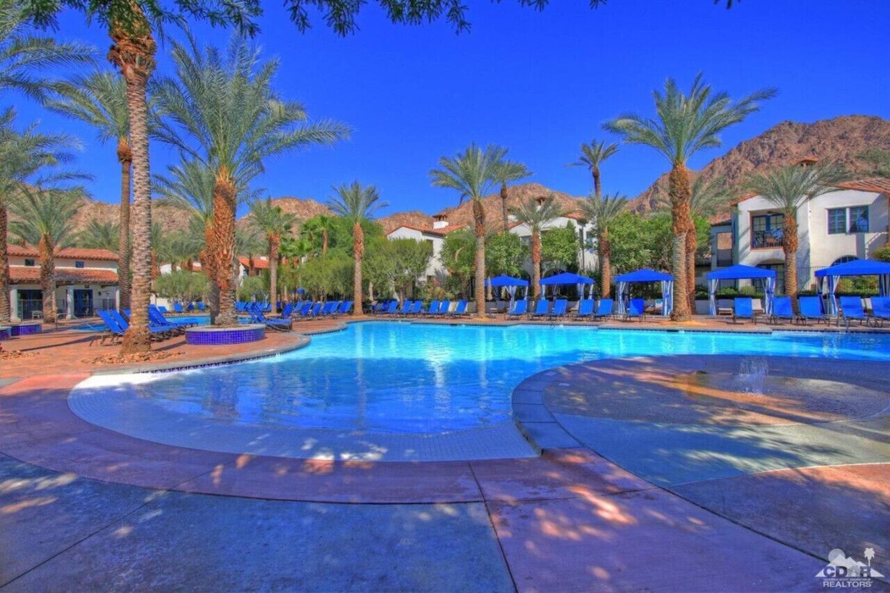 A swimming pool with blue lounge chairs and umbrellas, surrounded by palm trees and buildings with red-tiled roofs.