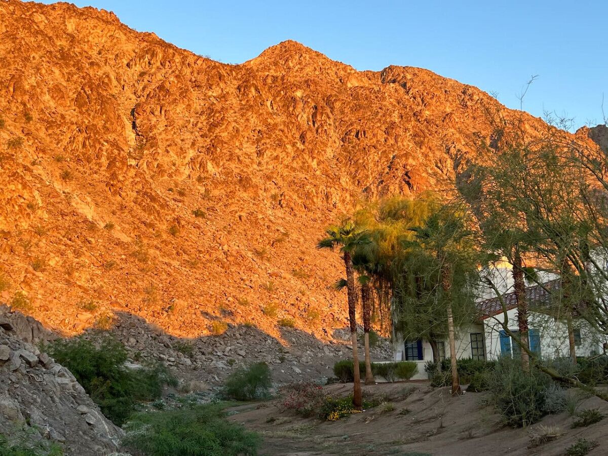 Paisaje desértico con una montaña al fondo y un edificio de fachada blanca y detalles azules.