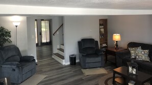 Ein Wohnzimmer mit einem dunkelgrauen Sofa, zwei Sesseln, einem Couchtisch und einem Bücherregal. Eine Treppe führt in einen anderen Raum.