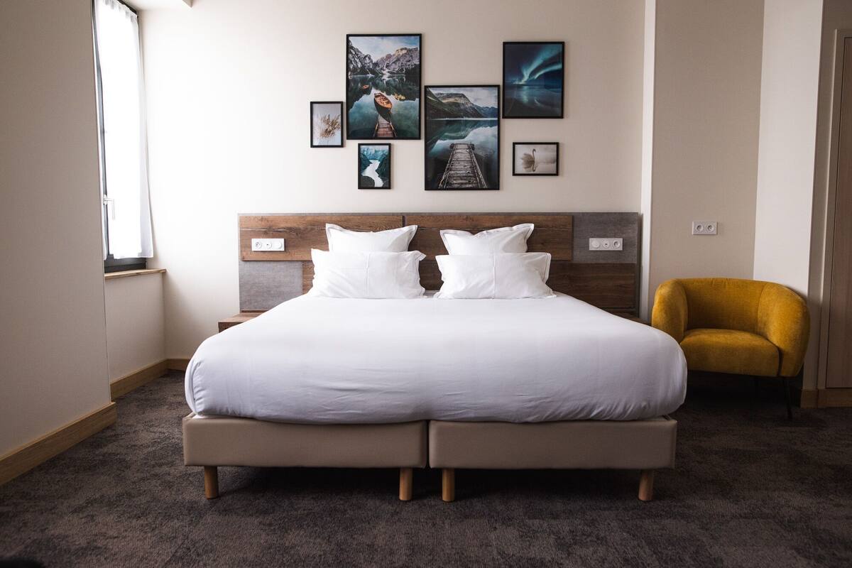 Uma cama grande com cabeceira de madeira, várias almofadas e uma poltrona amarela pequena num quarto de hotel.