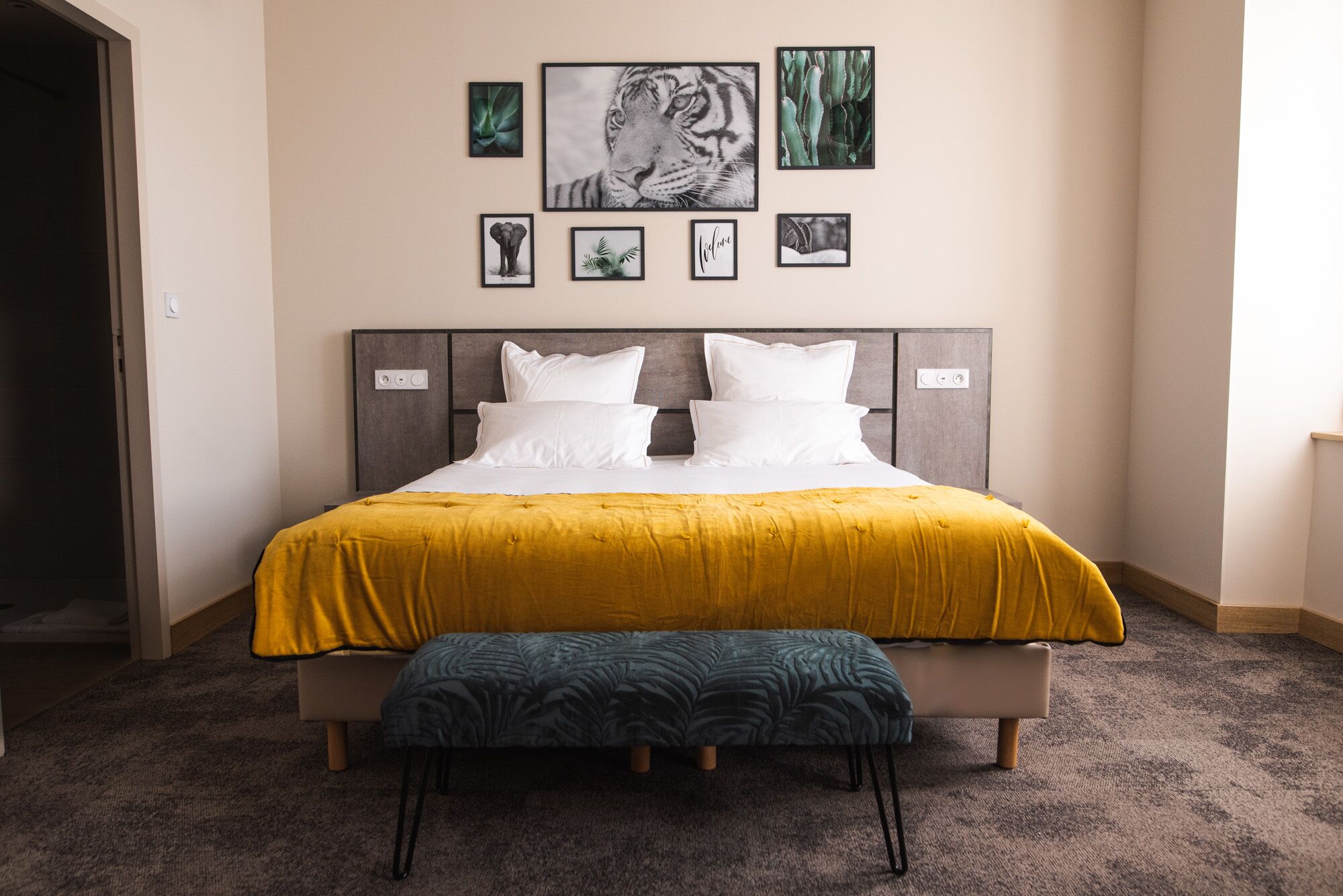 Una cama bien tendida con un edredón amarillo y un banco a juego, un cabecero con toma de corriente y varias fotografías enmarcadas en la pared sobre la cama.