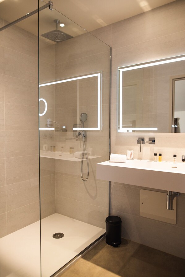 Un baño moderno con ducha de vidrio, un lavamanos blanco con espejo y un cesto de basura negro.