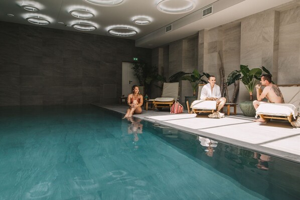 Un área de piscina interior moderna con una piscina cristalina, sillas de descanso e iluminación circular en el techo.