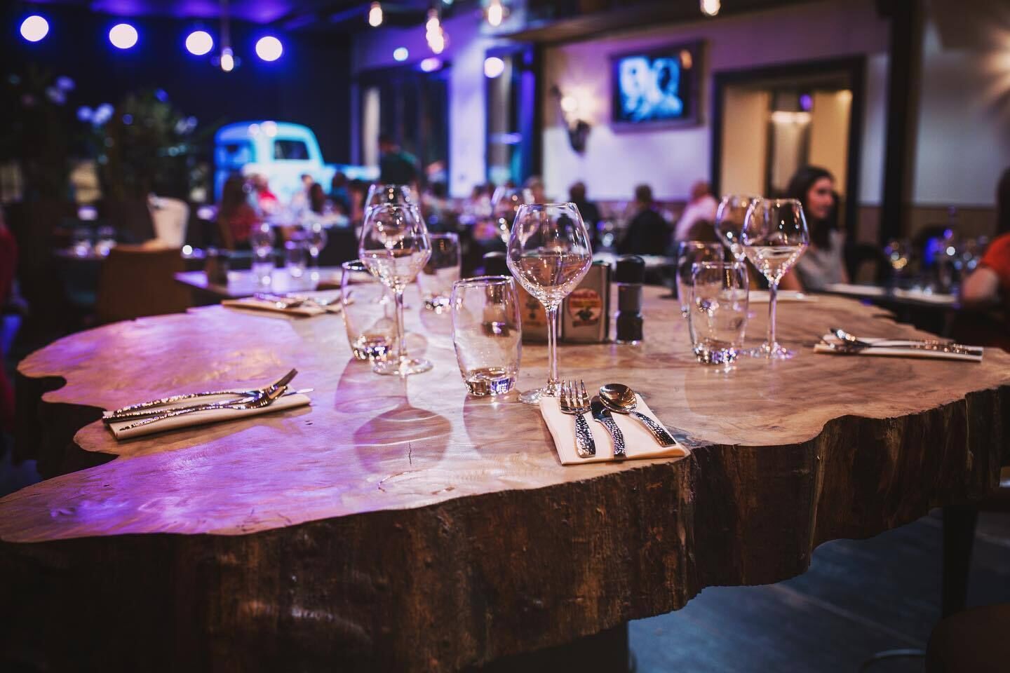 Een houten tafel met wijnglazen en bestek, in een schemerig verlicht restaurant met een televisiescherm op de achtergrond.