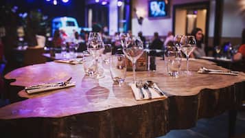 Una mesa de madera con copas de vino y cubiertos, en un restaurante con poca luz y una pantalla de televisión al fondo.