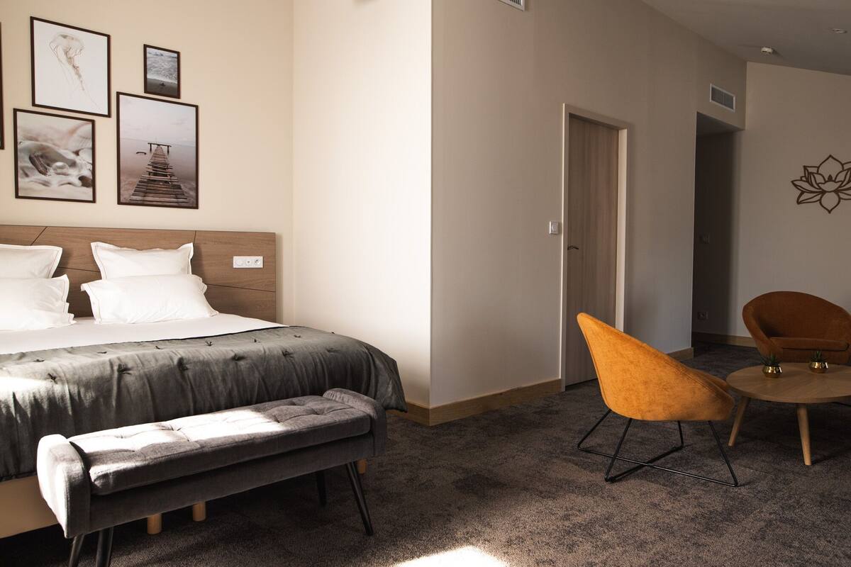 Quarto de hotel com cama, um banco, duas cadeiras e uma porta que dá para outro espaço.