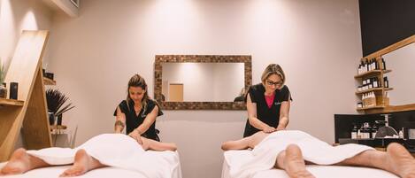 Ein Spa-Raum mit zwei Massagetischen, einem großen Spiegel und Regalen mit Flaschen.