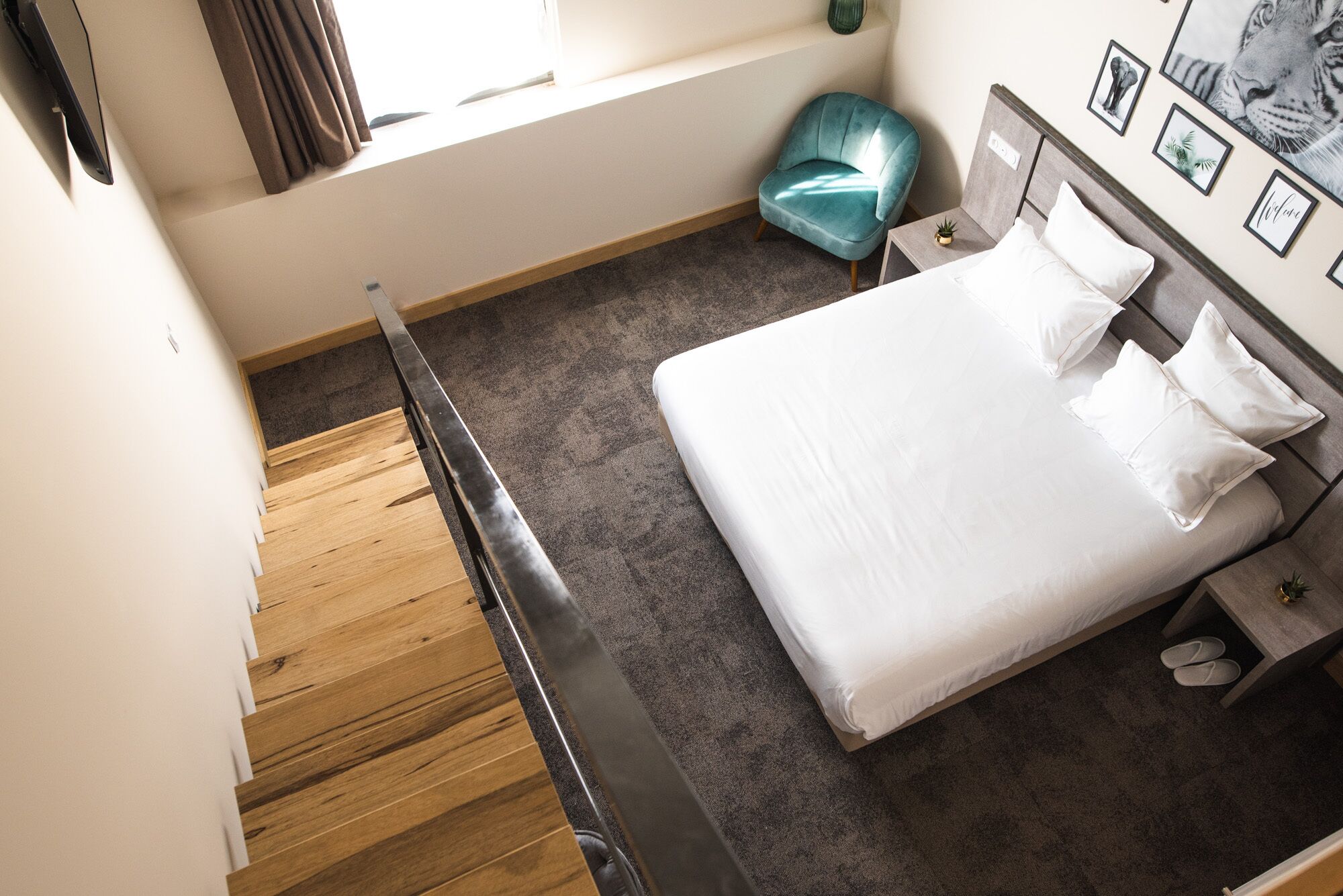 Habitación de hotel moderna con una escalera de madera, una cama con ropa blanca, un sillón color turquesa y una mesita con una planta.
