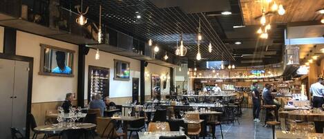 A spacious restaurant with multiple dining tables and chairs, hanging light fixtures, and a bar area in the background.